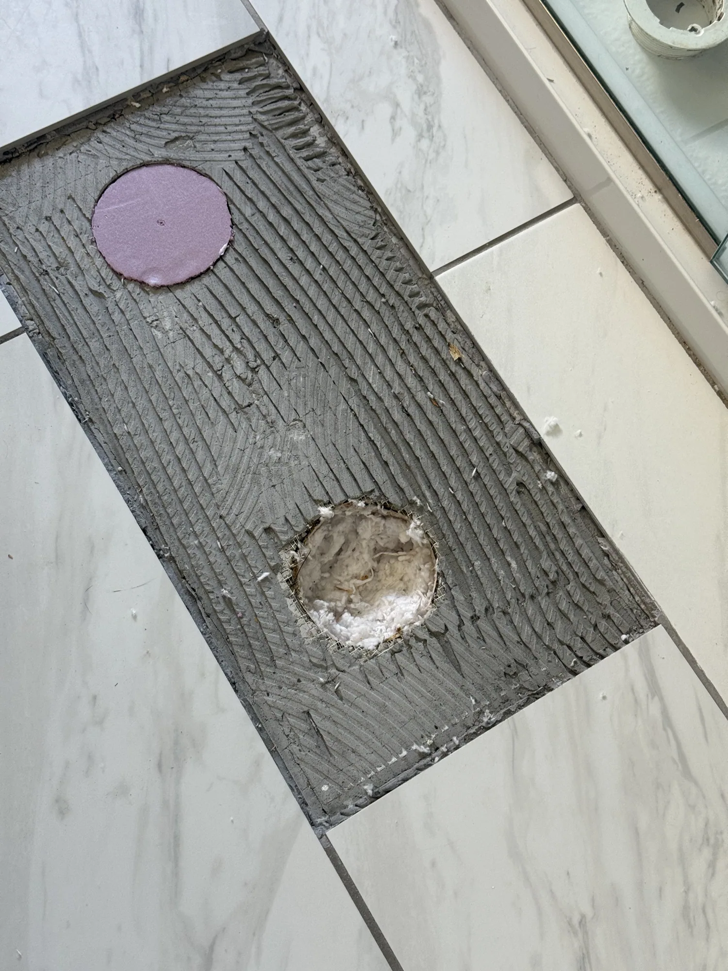 Cut backer board patch on a tile floor, one hole capped with a pink patch and one open with insulation packed around the pipe