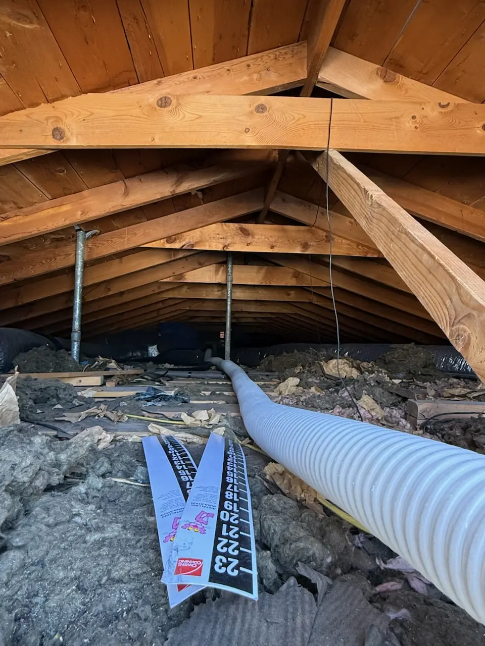 Attic with patchy older insulation, a flexible hose running across the joists and a depth marker card laid on top