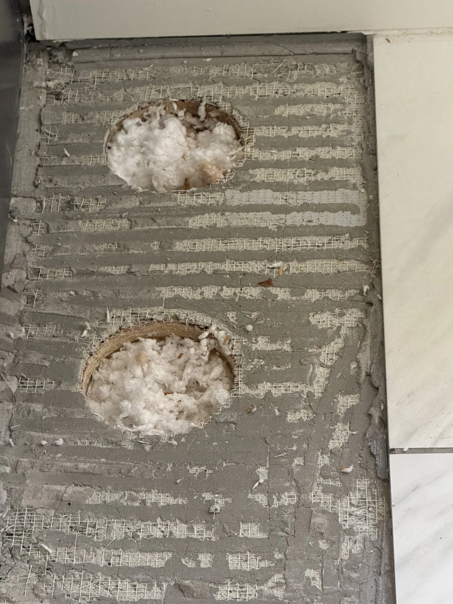 Two round holes cut in cement backer board on a tile floor, each packed with insulation around a pipe